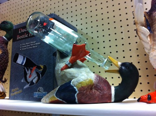 A decorative duck bottle holder with a glass bottle balanced on its beak, displayed on a shelf.