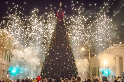 A large Christmas tree adorned with decorations, surrounded by fireworks and a crowd celebrating at night.