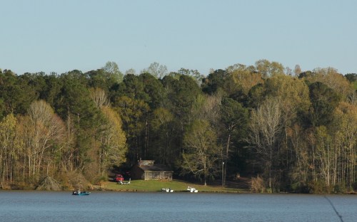 A serene lake scene with a cabin and boats, surrounded by lush trees and clear blue skies.