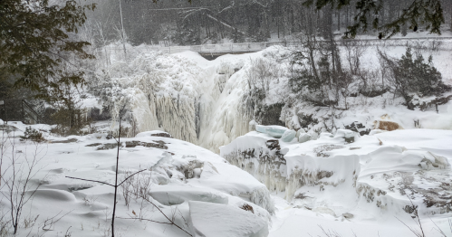 A frozen waterfall surrounded by snow-covered rocks and trees, with a bridge visible in the background.