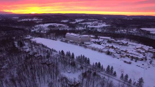 Aerial view of a snowy landscape at sunset, featuring a large building surrounded by trees and hills.
