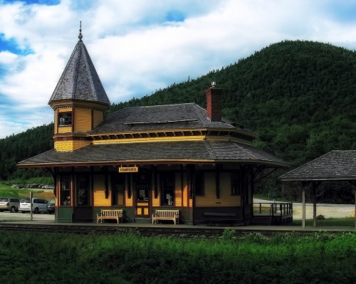 Historic train station with a pointed tower, surrounded by green hills and a blue sky.