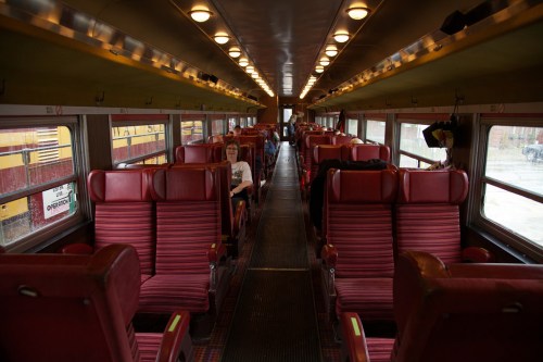 Interior of a train car with red upholstered seats and a few passengers, featuring overhead lights and large windows.