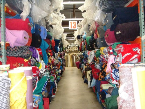 A colorful fabric store aisle lined with rolls of various textiles on both sides, creating a vibrant and busy atmosphere.