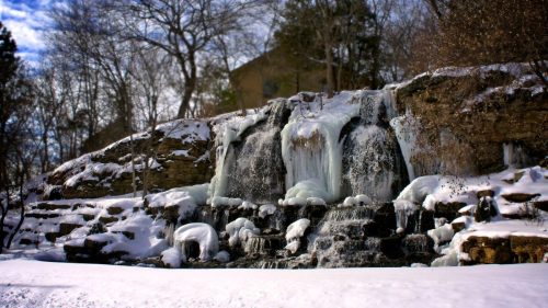 A frozen waterfall cascading over rocky terrain, surrounded by snow and bare trees under a cloudy sky.