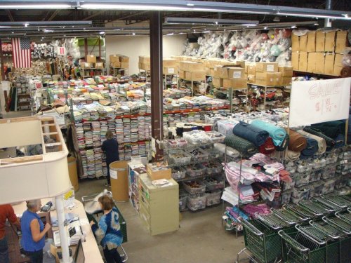 A large fabric store filled with stacked materials, shelves, and shoppers at the checkout area.