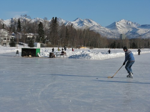 A person skating on an ice rink with mountains in the background, while others enjoy winter activities nearby.