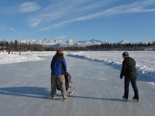 Two people skate on an ice path surrounded by snow and mountains under a clear blue sky.