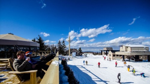 A snowy ski resort scene with people skiing and relaxing on a deck, enjoying the sunny winter day.