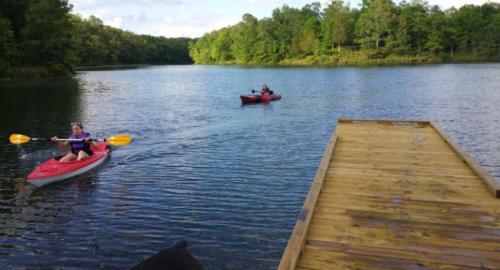 Two people kayaking on a calm lake, with a wooden dock in the foreground and lush greenery in the background.