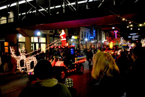 A festive parade scene with a decorated vehicle featuring Santa and reindeer, surrounded by onlookers and holiday lights.