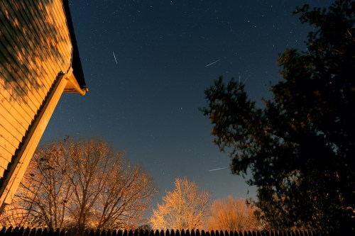 A night sky filled with stars and a few shooting stars, framed by trees and the side of a house.