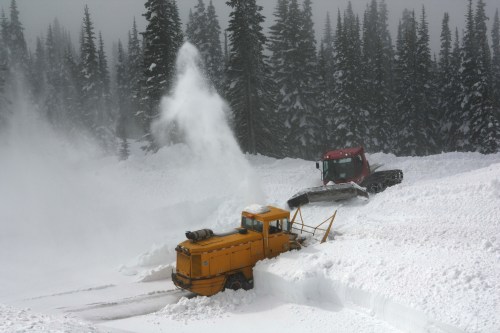 A yellow snowplow clears deep snow in a forested area, with a red machine assisting in the background.