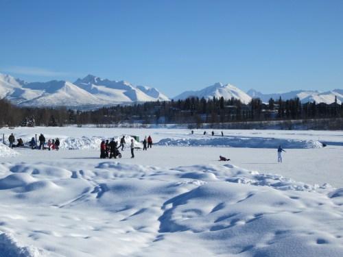 A snowy landscape with people ice skating and playing on a frozen lake, surrounded by mountains under a clear blue sky.