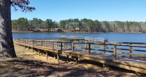A wooden pier extends into a calm lake, surrounded by trees under a clear blue sky.