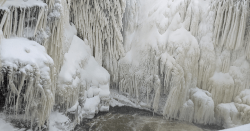 A frozen waterfall surrounded by icy formations and snow, with a river flowing at the base.