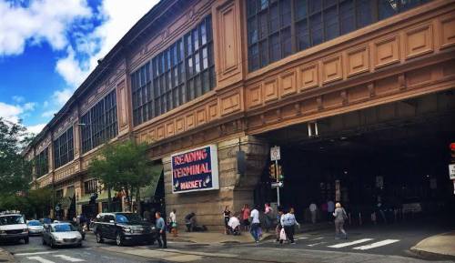 A bustling street scene outside Reading Terminal Market, featuring pedestrians and vehicles near a historic building.