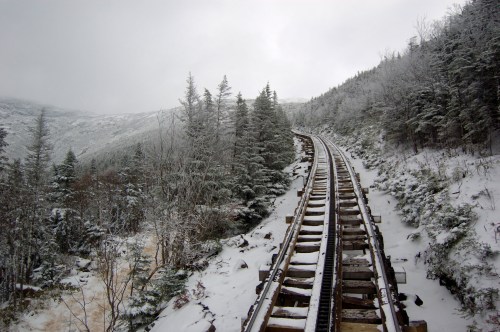 Snow-covered train tracks winding through a forest of evergreen trees in a mountainous landscape.