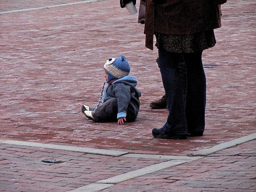 A toddler in a blue hat sits on a brick pavement, with an adult standing nearby.