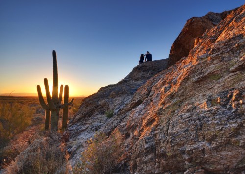A couple sits on a rocky ledge at sunset, with a tall cactus silhouetted against the colorful sky.