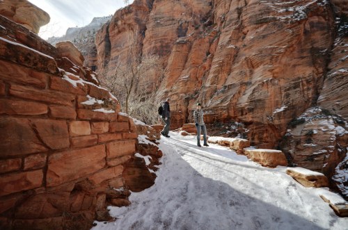 Two hikers on a snowy path surrounded by red rock formations in a canyon.