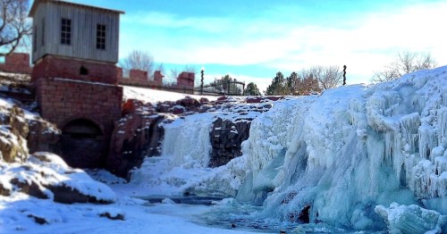 Frozen waterfall with ice formations, a historic building in the background, and a clear blue sky above.