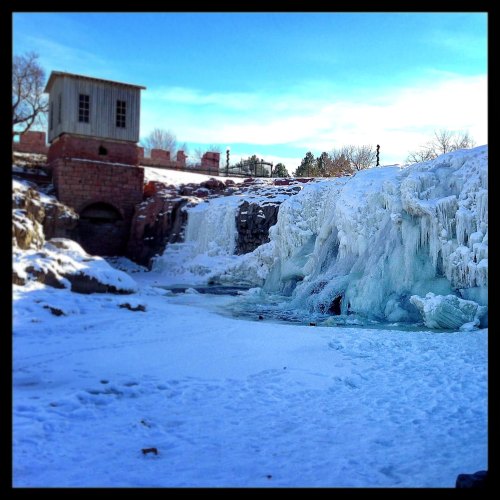 A frozen waterfall with ice formations, surrounded by snow, and an old building in the background under a clear blue sky.
