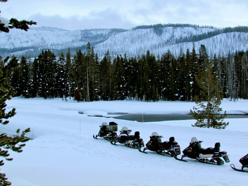 A line of snowmobiles parked on a snowy landscape, surrounded by trees and mountains under a cloudy sky.