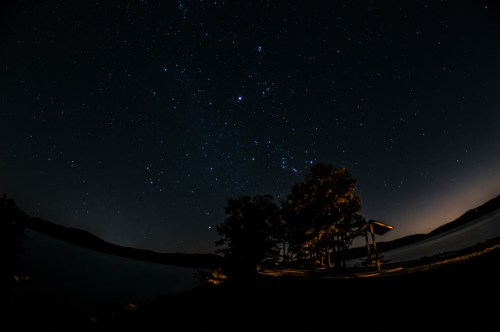 Starry night sky over a calm lake, with silhouettes of trees and a gazebo illuminated by soft light.