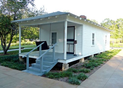 A small white house with a porch, steps, and a sign, surrounded by greenery and a paved walkway.