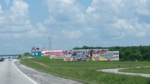 Billboard advertising a candy factory with free chocolate samples, snacks, and drinks, alongside a highway.