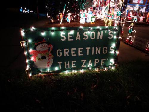 A festive sign reading "Season's Greetings The Zape's," illuminated with colorful lights and surrounded by holiday decorations.