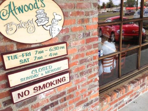 Sign for Atwood's Bakery with hours, closed Sunday and Monday, and a "No Smoking" notice, next to a window view.