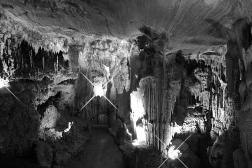 A dramatic black and white image of a cave with stalactites and stalagmites illuminated by soft lighting.