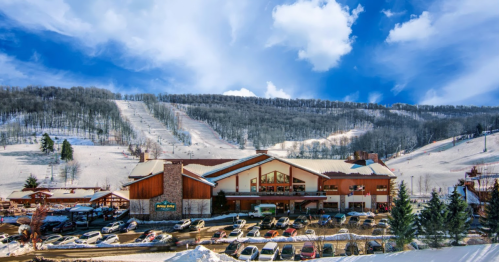 A ski resort building with a parking lot, surrounded by snow-covered slopes and trees under a blue sky.