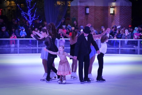 A group of skaters performs on ice, with a young girl in a pink dress at the front, surrounded by an audience.