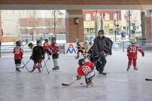 Children in hockey gear skate and practice on an ice rink, with a coach guiding them in a winter setting.