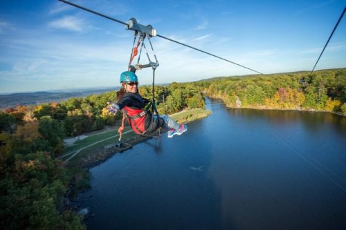 A person zip-lining over a scenic lake surrounded by colorful autumn trees under a clear blue sky.
