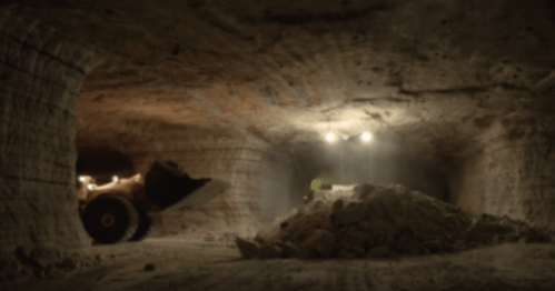 An underground mining tunnel with machinery and a pile of extracted material, illuminated by overhead lights.