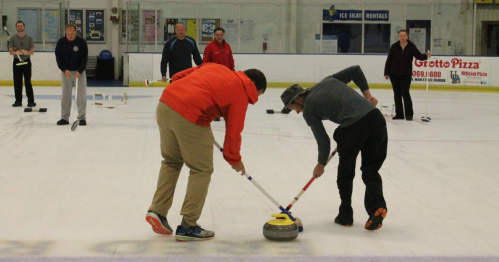 Two players curl a stone on ice while others watch in the background, engaged in a curling practice session.