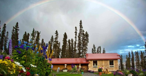 A cozy stone cabin surrounded by colorful flowers and tall trees, with a vibrant rainbow arching overhead.