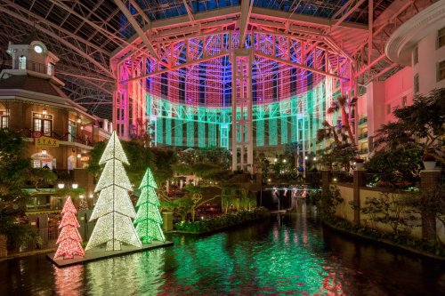 Colorful holiday lights illuminate a large indoor atrium with decorated trees and a water feature.