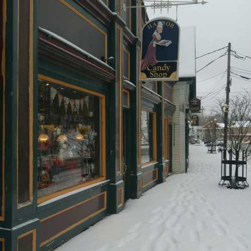 A quaint candy shop with a sign, surrounded by snow-covered streets and trees, creating a cozy winter scene.