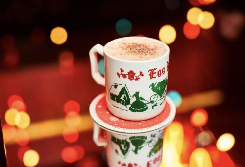 A festive mug of eggnog sits on a decorative saucer, surrounded by colorful bokeh lights.