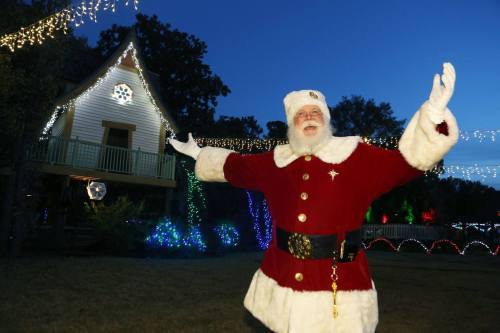 Santa Claus stands with open arms in front of a beautifully lit house, surrounded by festive holiday decorations.