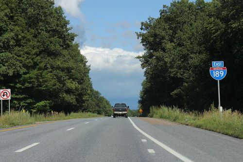 A highway scene with a truck driving on a road, flanked by trees and an Interstate 189 East sign.