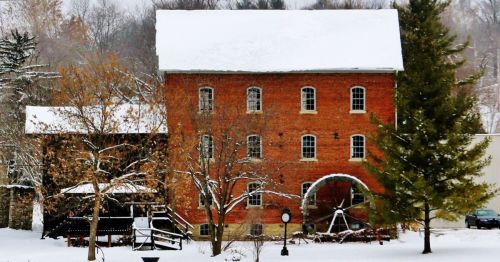 Historic brick building with a water wheel, surrounded by snow-covered trees and a gazebo in a winter landscape.