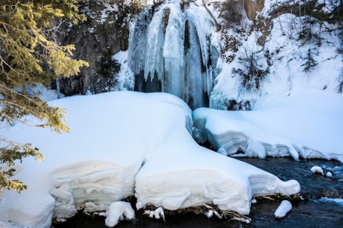 A frozen waterfall surrounded by snow-covered ground and icy formations, with trees in the background.