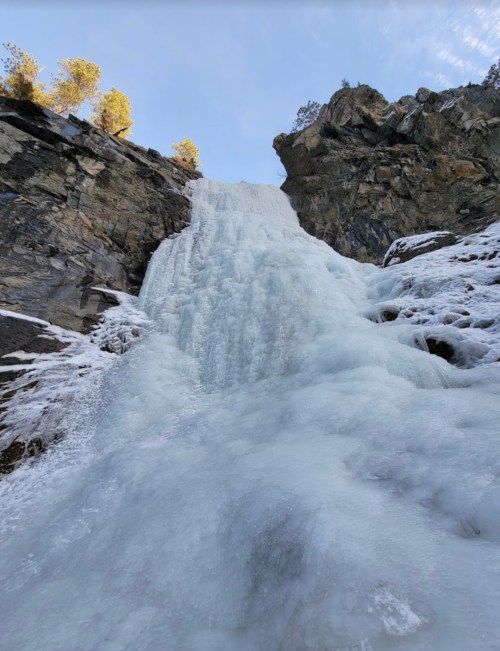 A frozen waterfall cascading down rocky cliffs, surrounded by snow and trees under a clear blue sky.