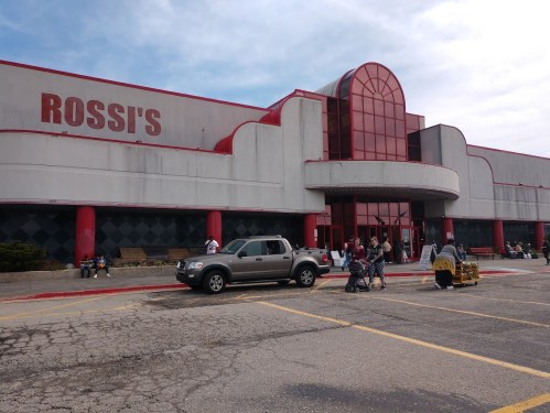 Exterior of Rossi's store with a red and gray facade, featuring shoppers and vehicles in the parking lot.